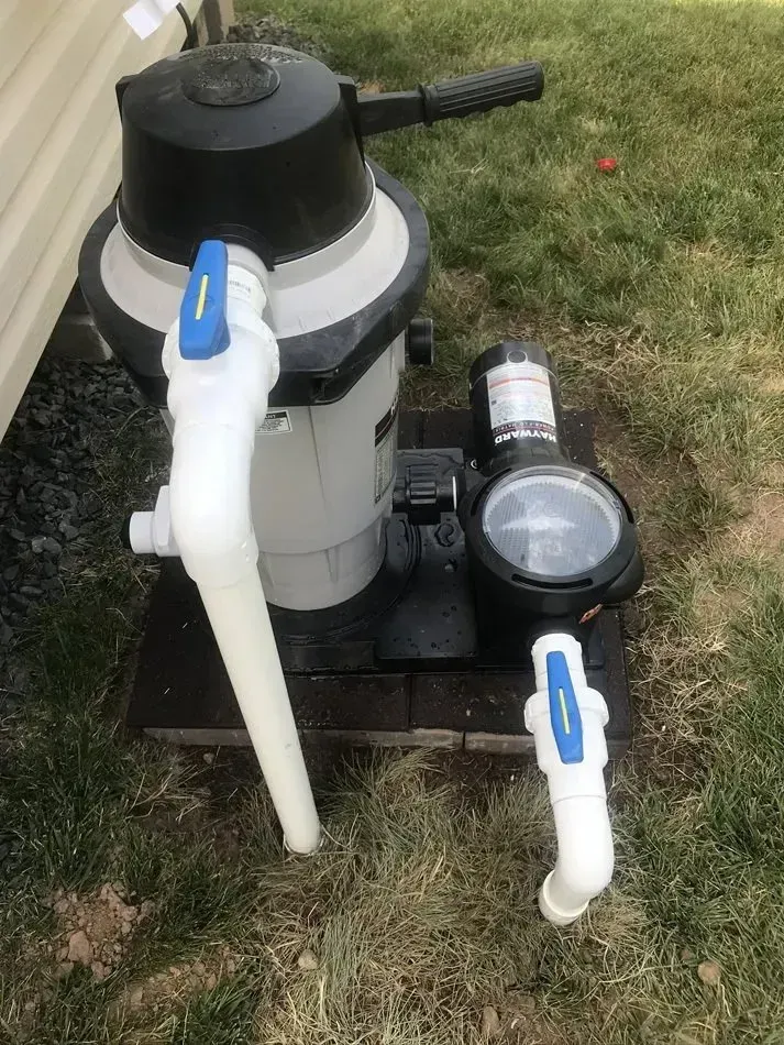 Pool pump and filter system with white pipes and blue-handled valves beside a house exterior.