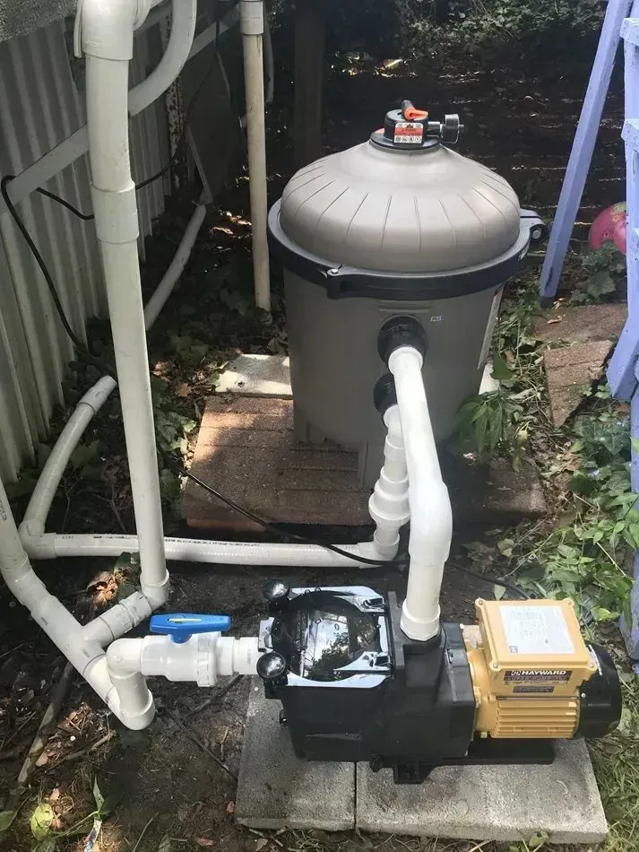 Pool equipment: gray filter tank, pump, and white PVC pipes beside a fence.