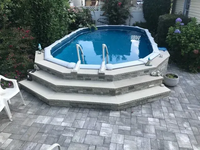 Backyard above-ground pool with stone steps, white railings, and a gray paver patio surrounded by greenery