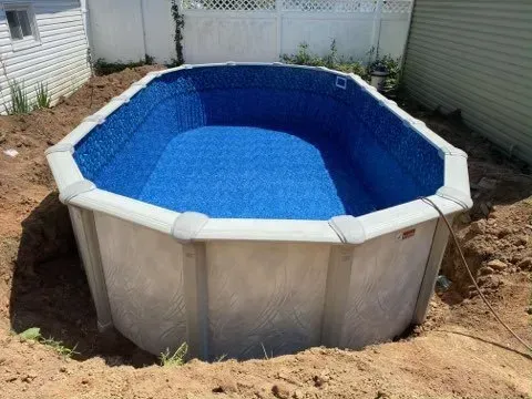 Above-ground pool with blue water in a backyard, surrounded by dirt and white fencing.