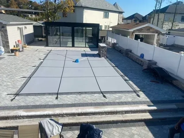 Backyard pool covered with a gray safety cover, surrounded by stone decking and a house in the background