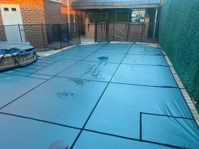 Blue-covered backyard patio with a car under a tarp, brick house, and metal gate in the background