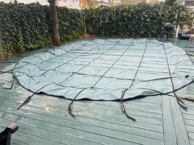 Covered backyard deck with a gray tarp secured by straps, bordered by hedges and trees.