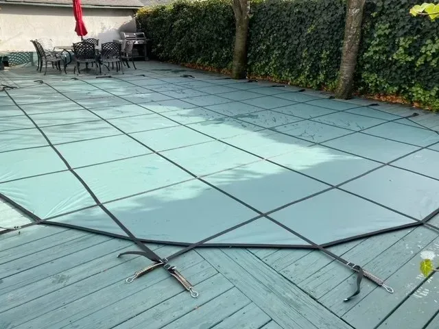 Patio covered with a taut blue pool safety cover, bordered by hedge and outdoor seating
