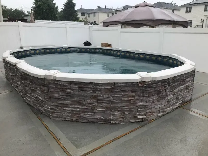 Round above-ground pool with stone-pattern exterior on a concrete patio in a fenced backyard