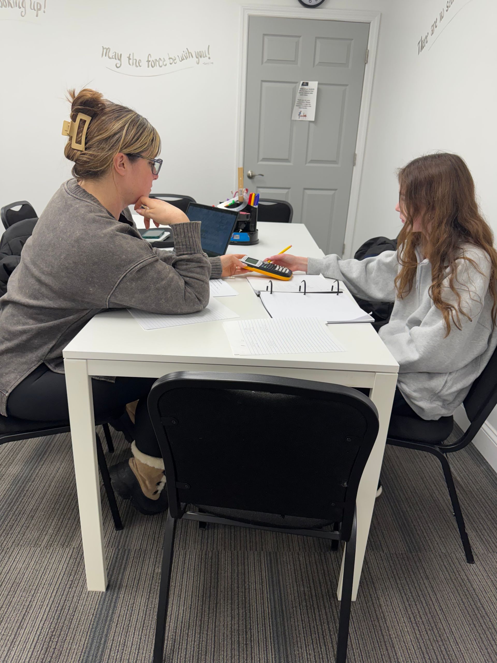 Two people at a white table, one pointing at papers.  A notebook and laptop are on the table. A door is in the background.