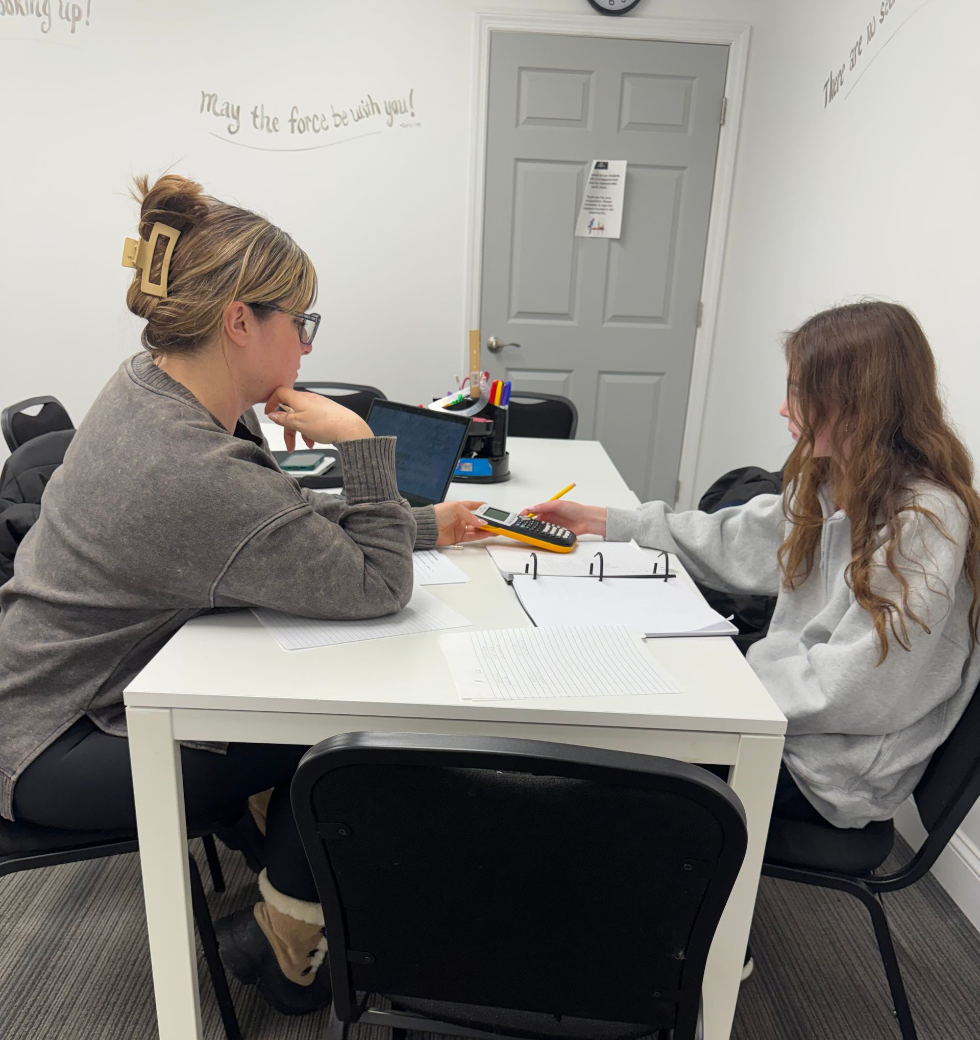 Two people at a white table, one pointing at papers. A notebook and laptop are on the table. A door is in the background.