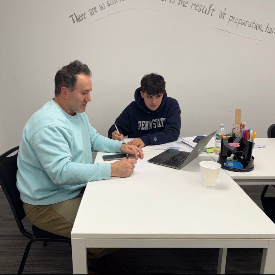 Two people sit at a white table in an office, working on documents together with a tablet present.