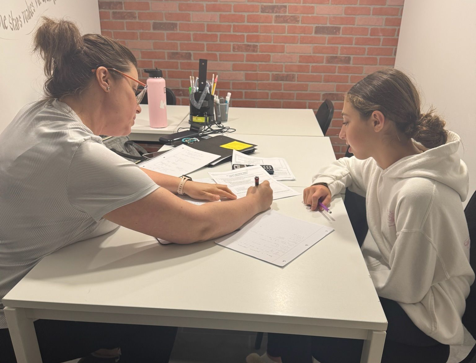 A person helps a young person seated at a table, both working on papers in a room.