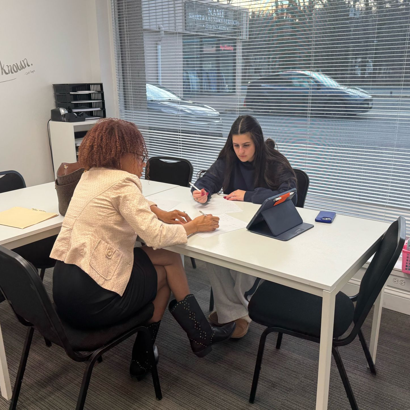 Two people sit at a white table in an office, working on documents together with a tablet present.