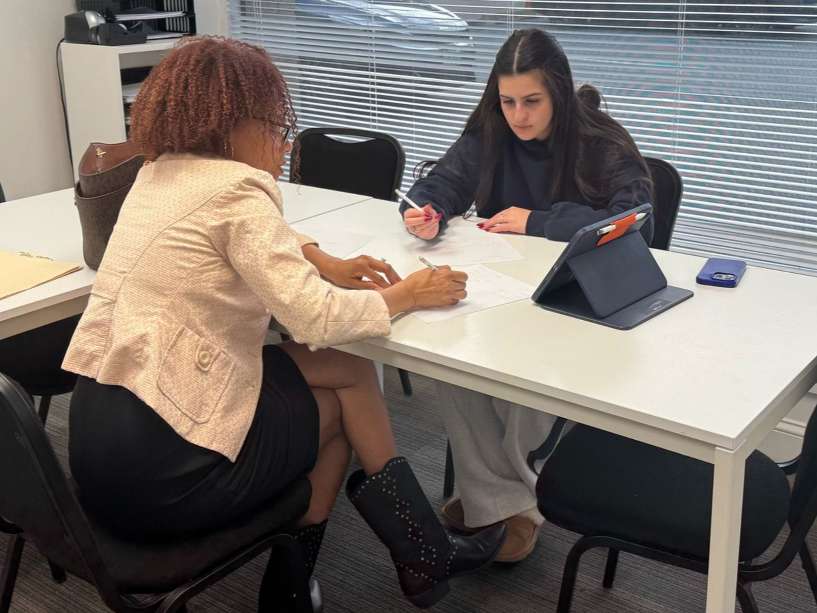 Two people sit at a white table in an office, working on documents together with a tablet present.