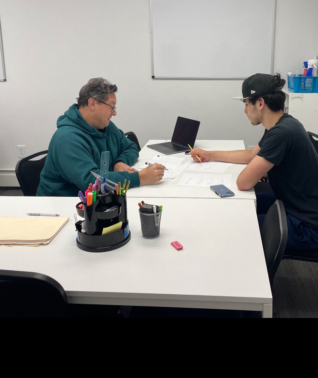 Two people sit at a white table in an office, working on documents together with a tablet present.