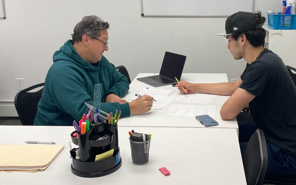 Two people sit at a white table in an office, working on documents together with a tablet present.