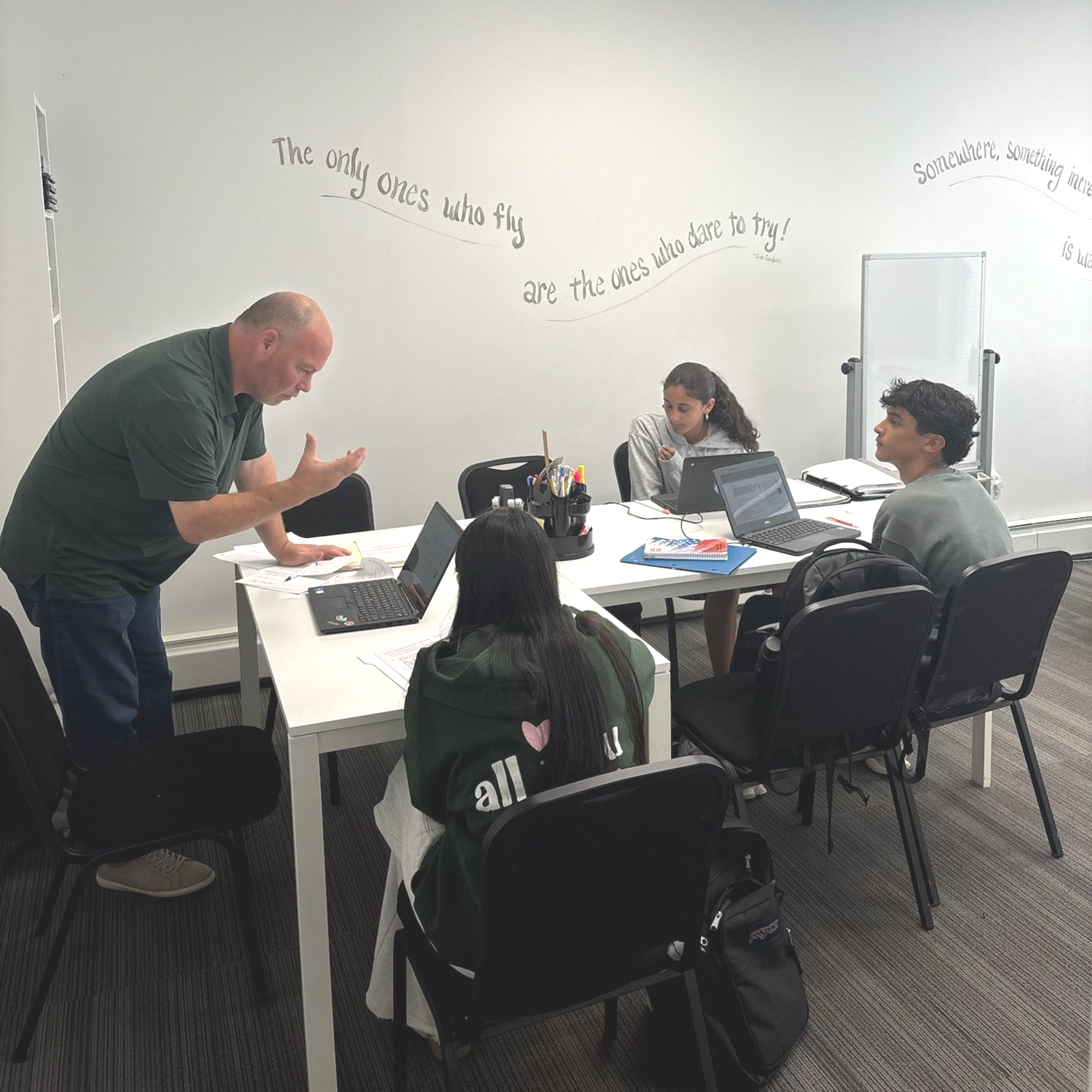 Teacher gestures while explaining to students seated at a table with laptops in a classroom.