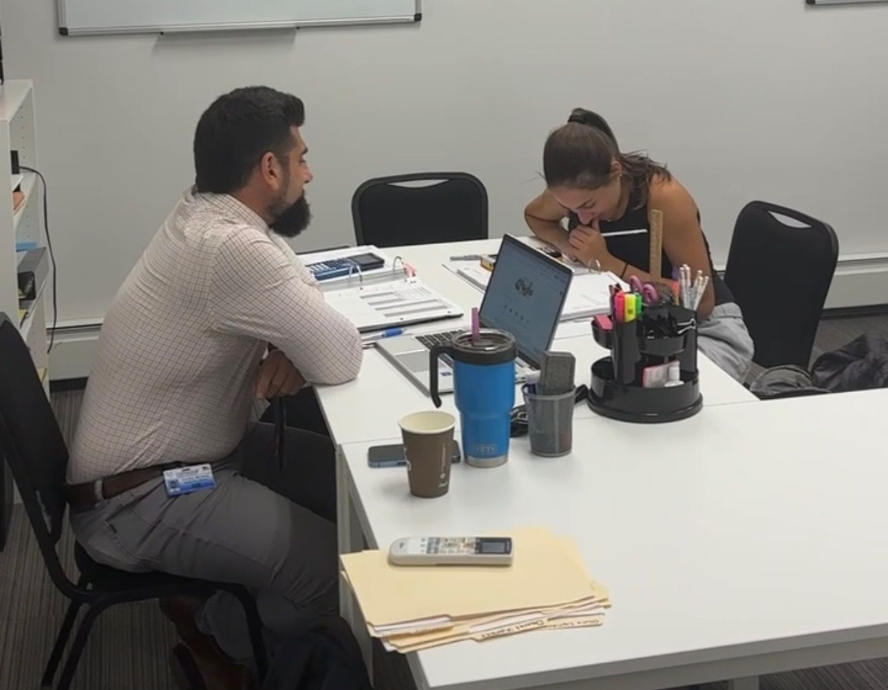 Two people at a desk: man looking at the woman writing on paper, laptop, coffee cups, and office supplies.