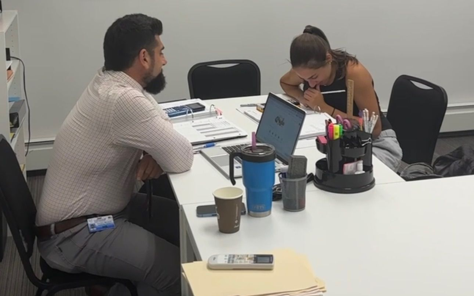 Two people at a desk: man looking at the woman writing on paper, laptop, coffee cups, and office supplies.