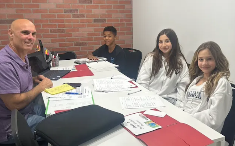 Four people sit around a table in a classroom. A man smiles at the camera, students are looking forward. Papers and a laptop are on the table.
