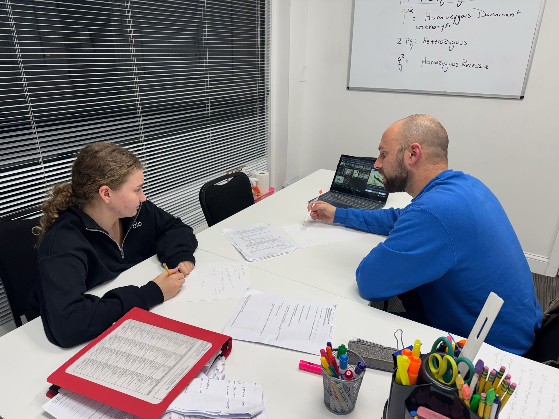 A person in a blue sweater sits at a table, instructing a student, both focused on papers in a bright, modern office.