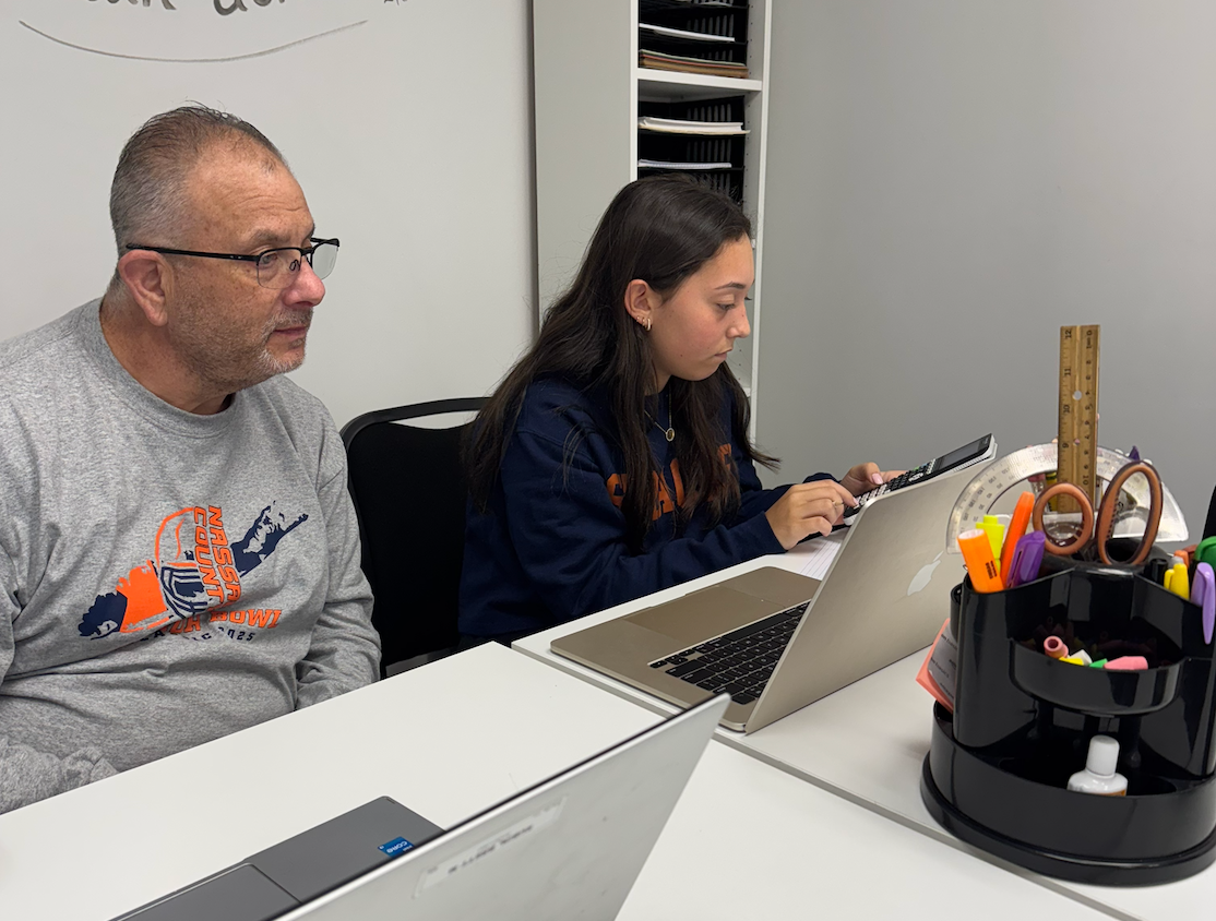 An adult and a student work side-by-side at a desk with laptops and school supplies in a bright classroom.