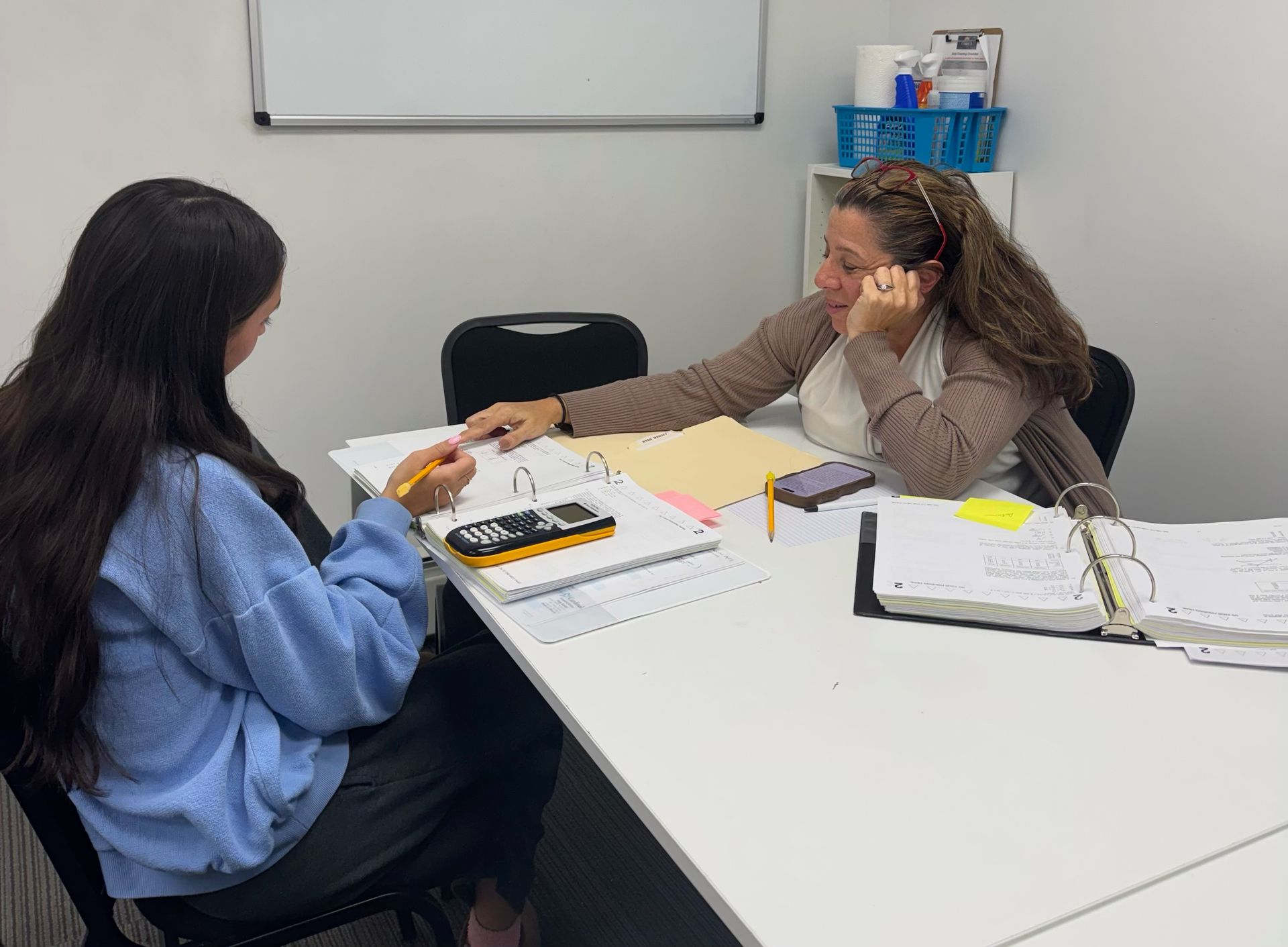 Two people sit at a desk in an office, reviewing documents and working with a calculator.