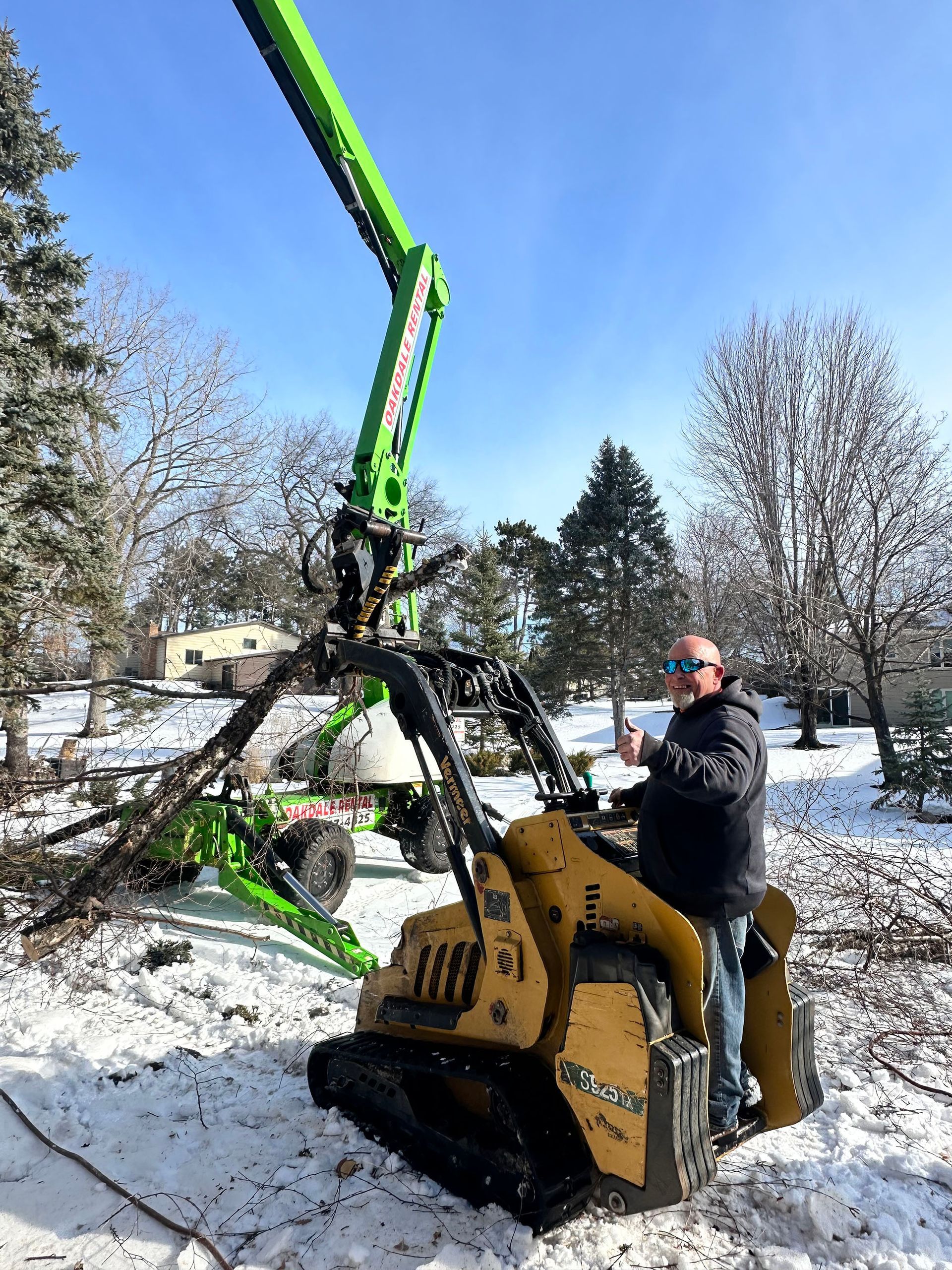 Person operating a green boom lift beside a yellow compact machine in a snowy outdoor yard