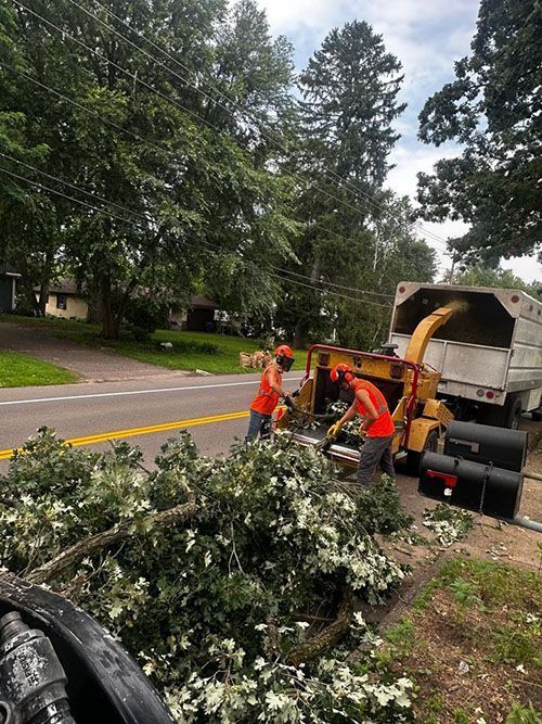 Workers clearing storm debris beside a street with a chipper truck and piles of branches