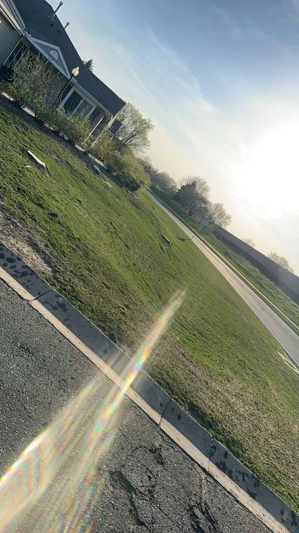 Tilted view of a grassy roadside field with a house edge, paved path, and bright sun flare in the sky
