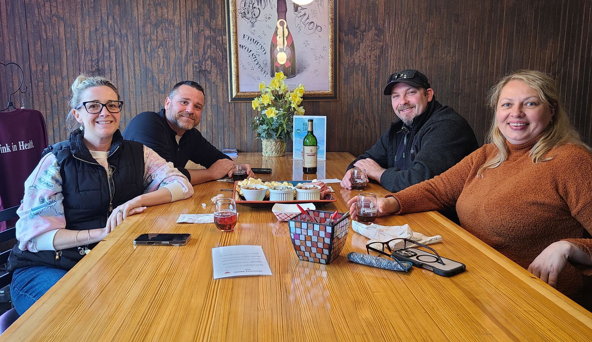 Four people seated around a wooden dining table in a cozy restaurant, smiling with drinks and snacks.