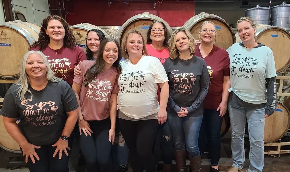 Group of people smiling indoors, posing in matching graphic shirts in front of wooden barrels.