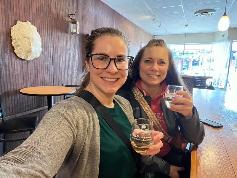 Two women smiling and toasting drinks in a cozy bar or café.