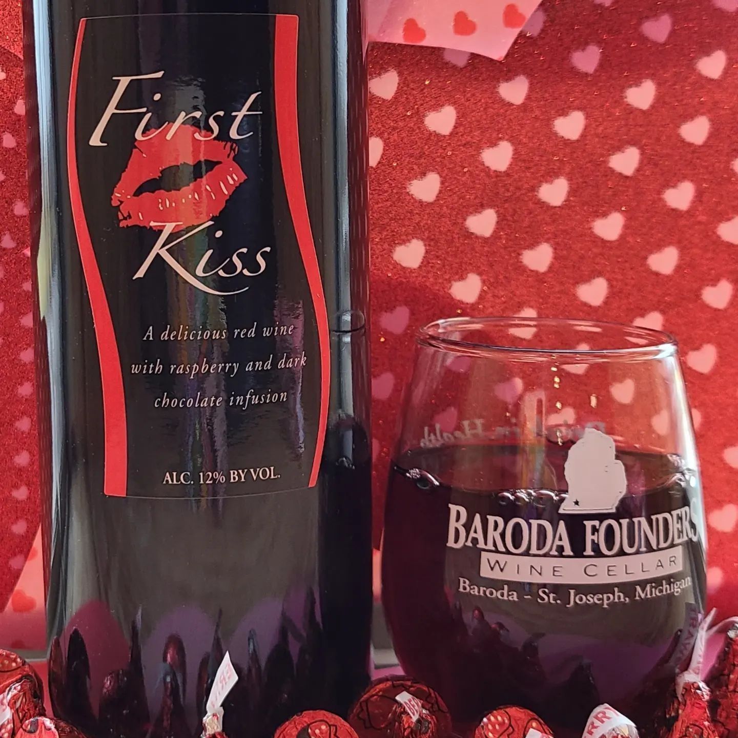 Wine bottle labeled “First Kiss” beside a Baroda Founders wine glass on a red heart-patterned background