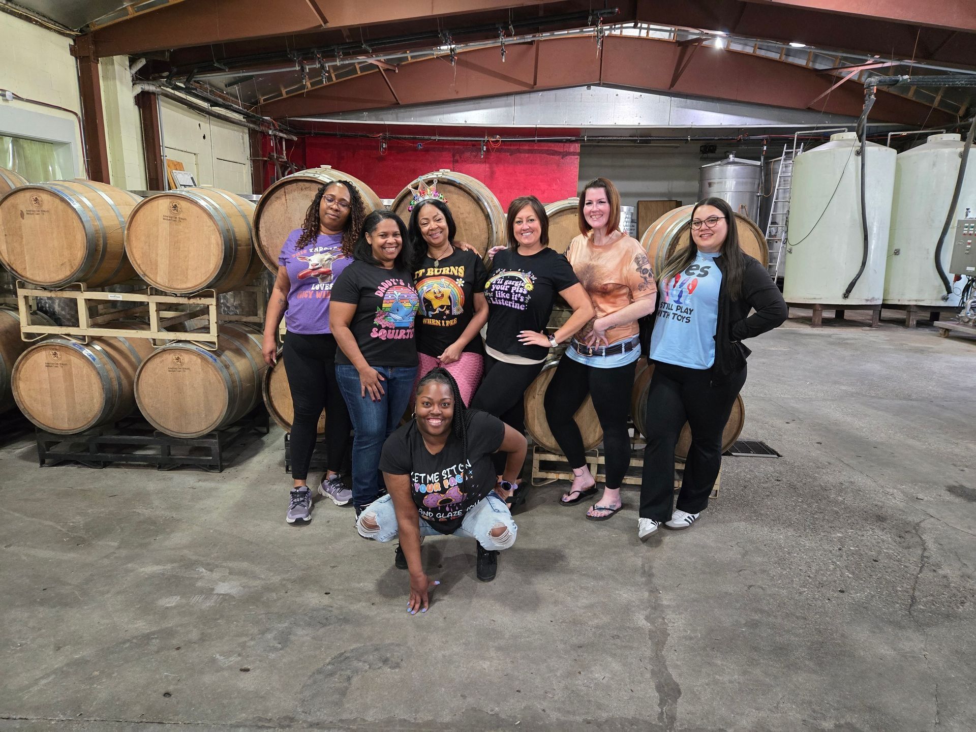 Group posing together in a winery barrel room, with large wooden barrels and a red wall backdrop.