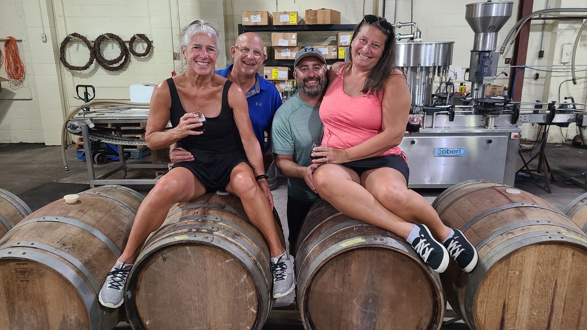 Four people sitting on wine barrels in a brewery, smiling and posing together
