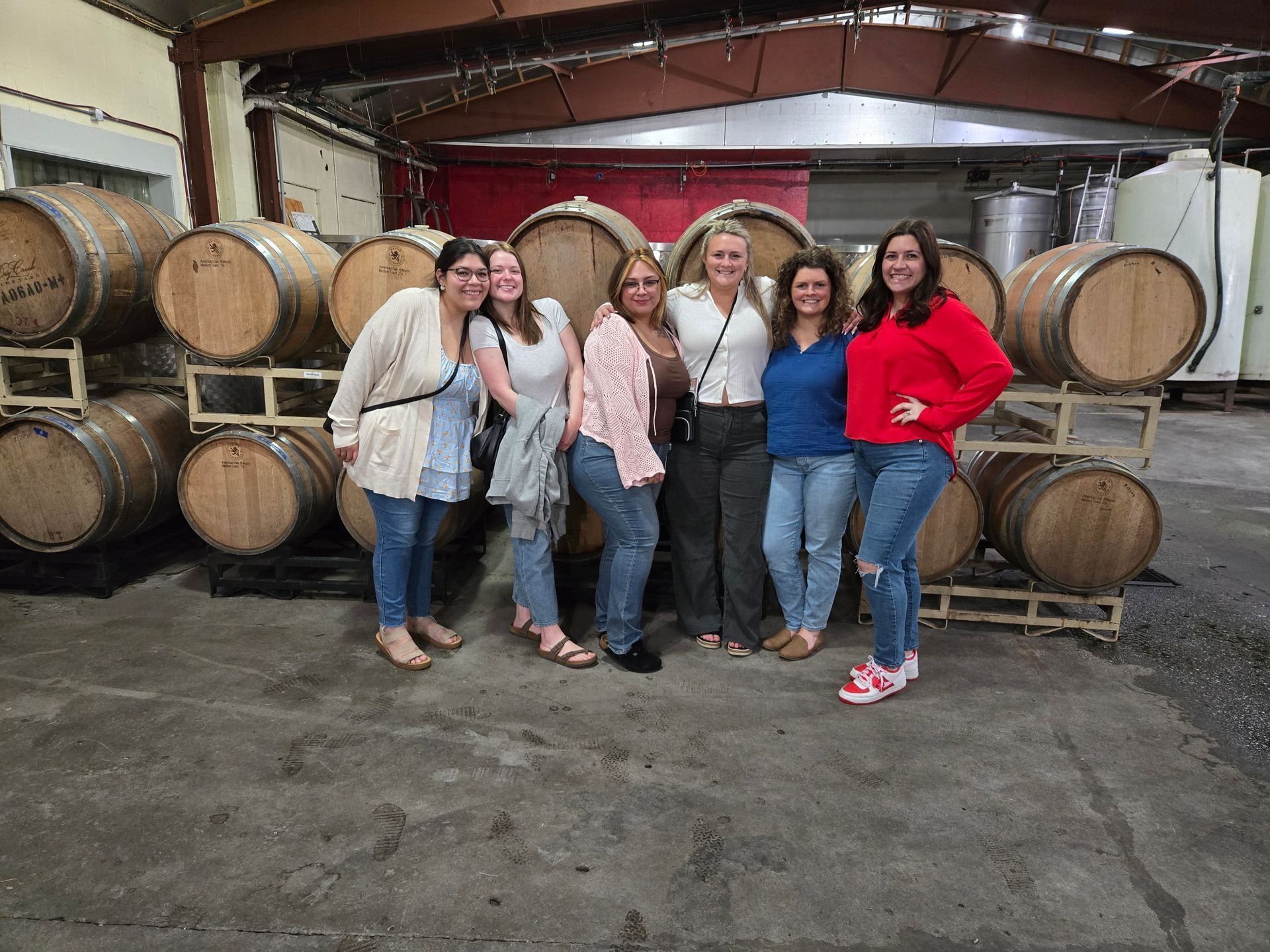 Six women posing in a winery cellar in front of stacked wooden barrels
