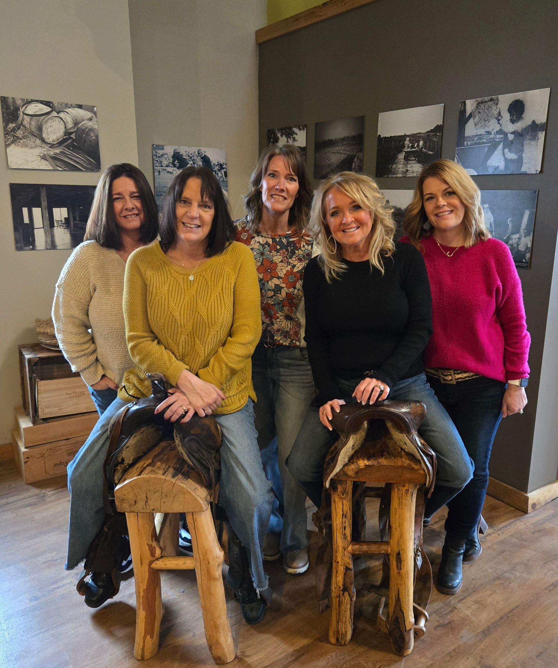 Five women seated on wooden stools in a casual indoor setting, smiling at the camera.