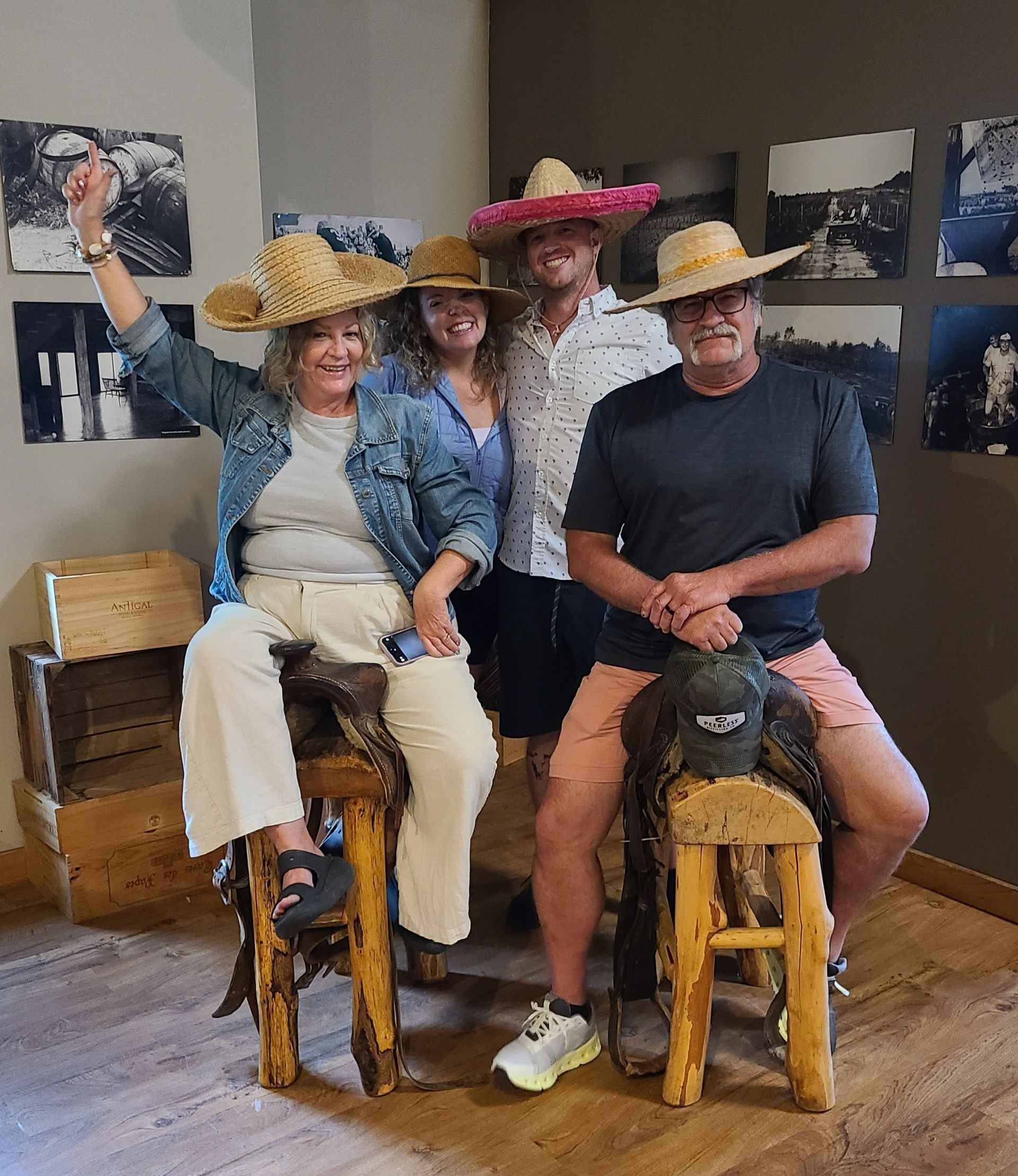 Three people wearing straw hats sit on wooden stools in an art gallery, posing with smiles.