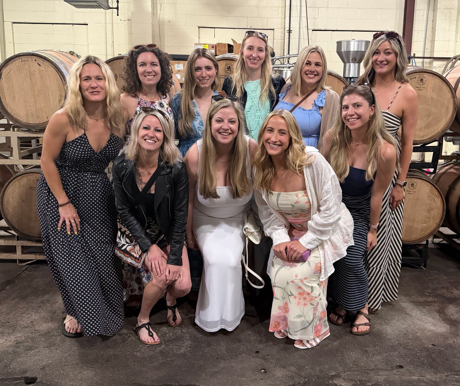 Eleven women pose in a barrel room, smiling in front of stacked wooden wine barrels.