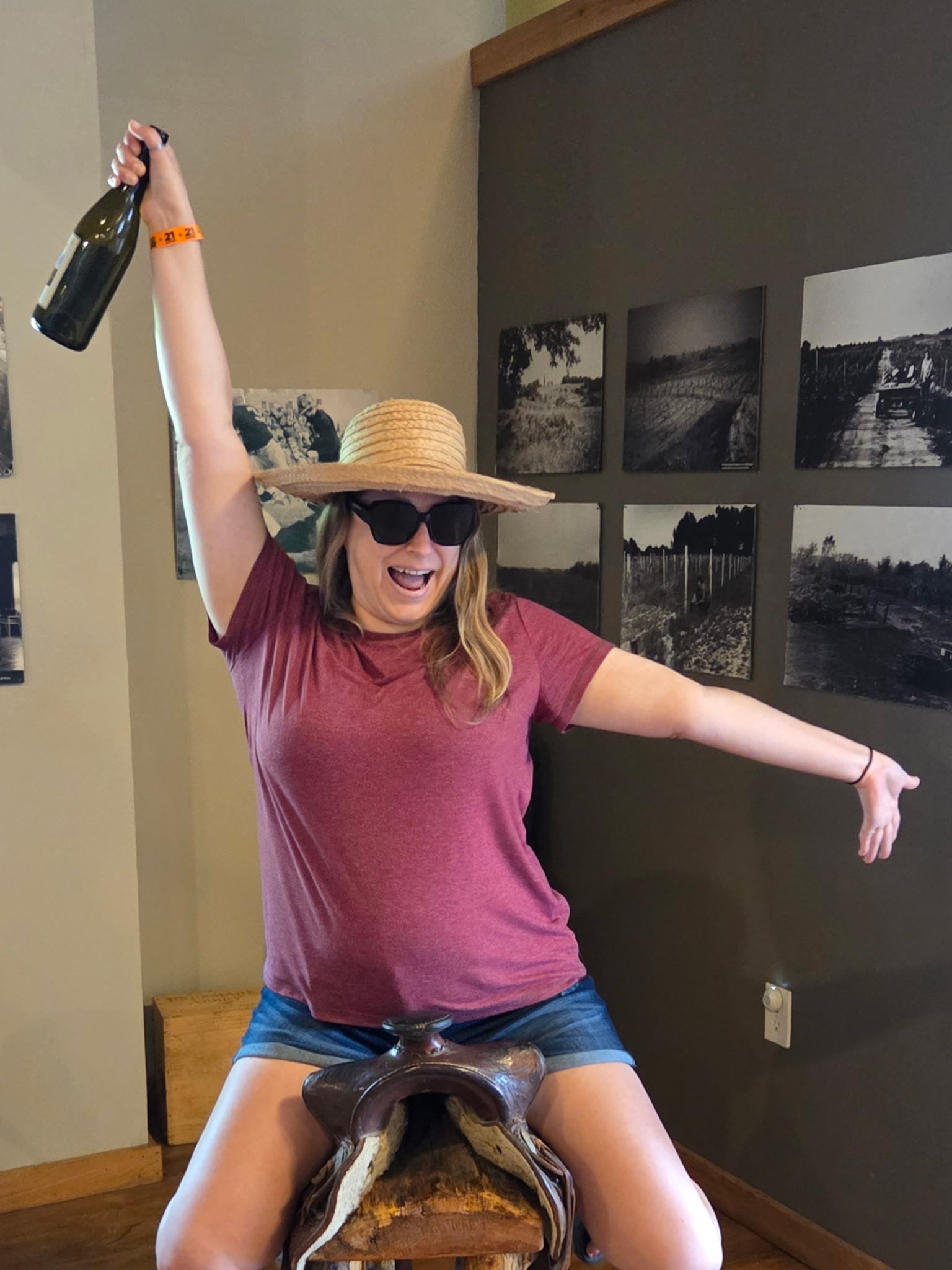 Smiling person in straw hat and sunglasses rides a mechanical bull, arm raised with a bottle.