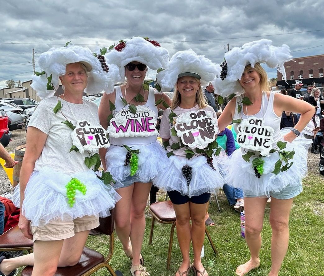 Four people in white feathered costumes and hats posing outdoors at a Pride event.