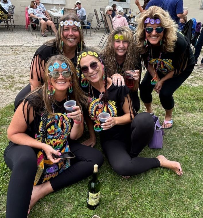 Five friends posing on grass with colorful face paint, sunglasses, and drinks at an outdoor party