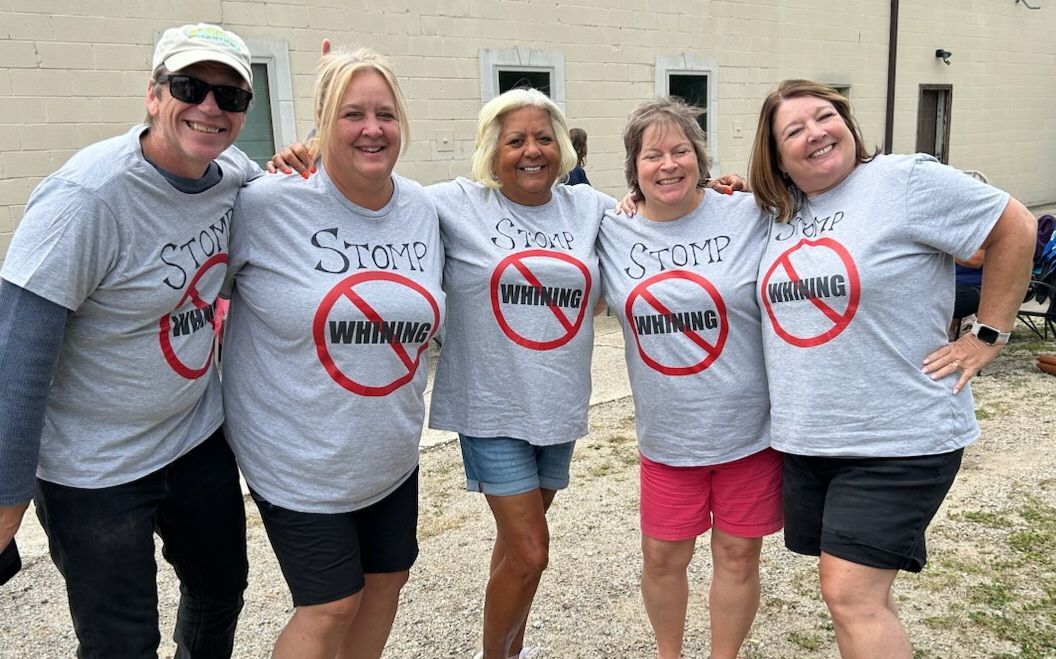 Five people in matching gray shirts stand smiling outdoors in front of a building.