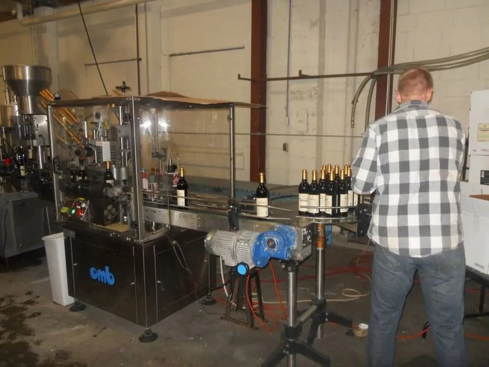 Bottling line in a warehouse, with a worker in a plaid shirt inspecting bottles on a conveyor.