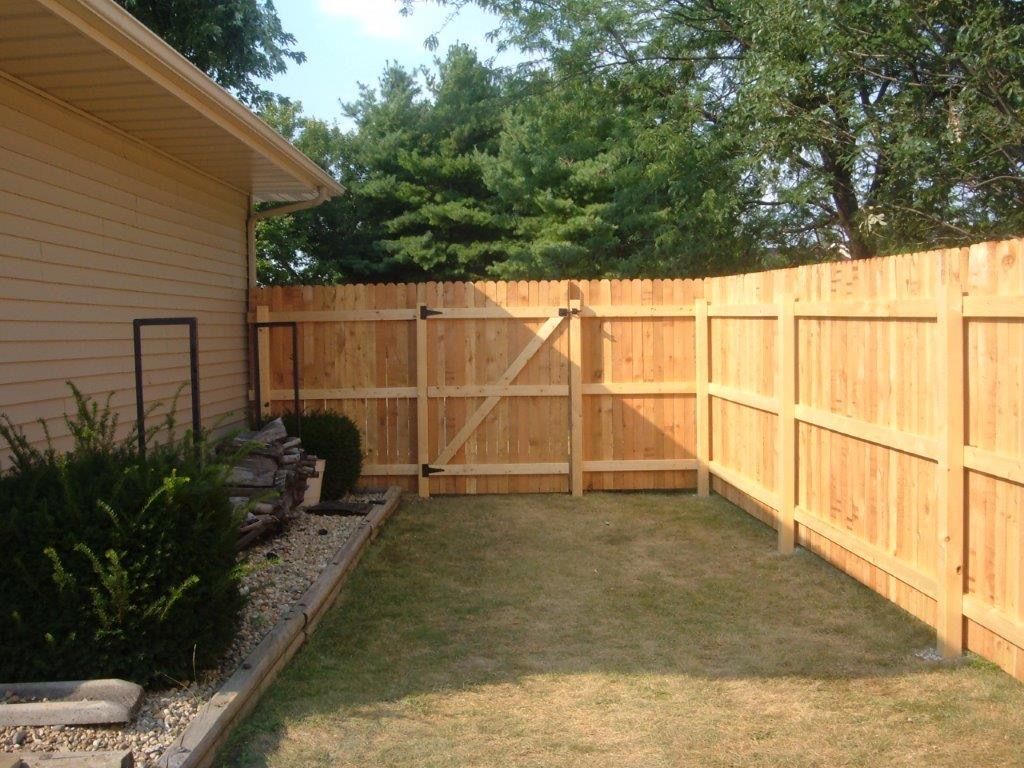 Wooden fence encloses a small backyard with grass and a gate; attached to a tan house.
