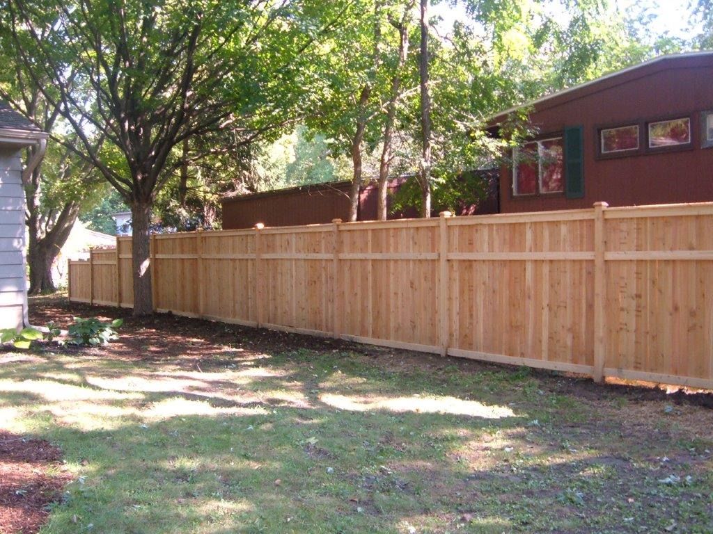 Wooden fence in a backyard, with a house and trees in the background. Sunny day.