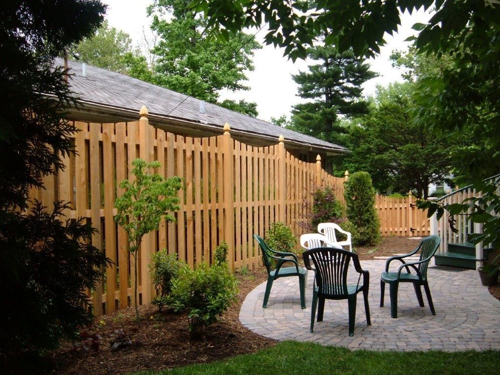 Wooden fence surrounds a patio with green chairs and a brick ground, nestled in a backyard with trees.