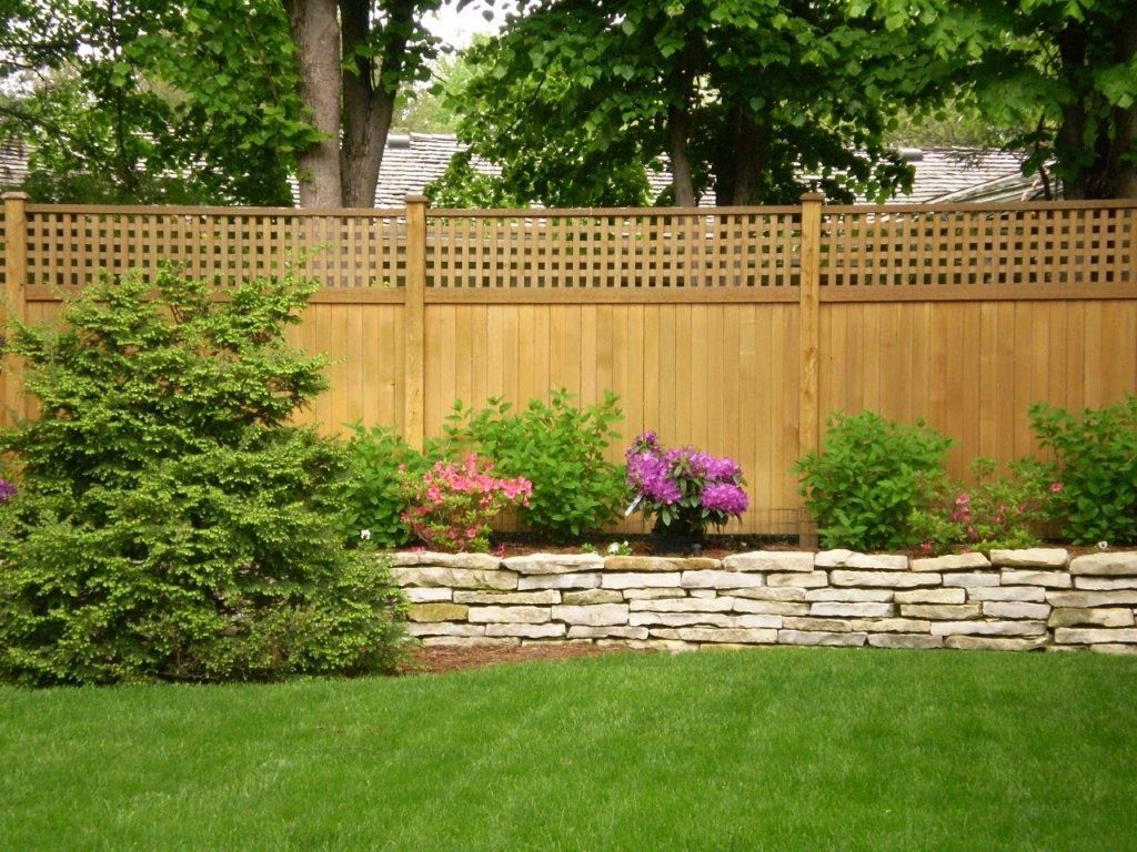 Wooden fence with lattice top, stone retaining wall, and green lawn with shrubs and flowers.