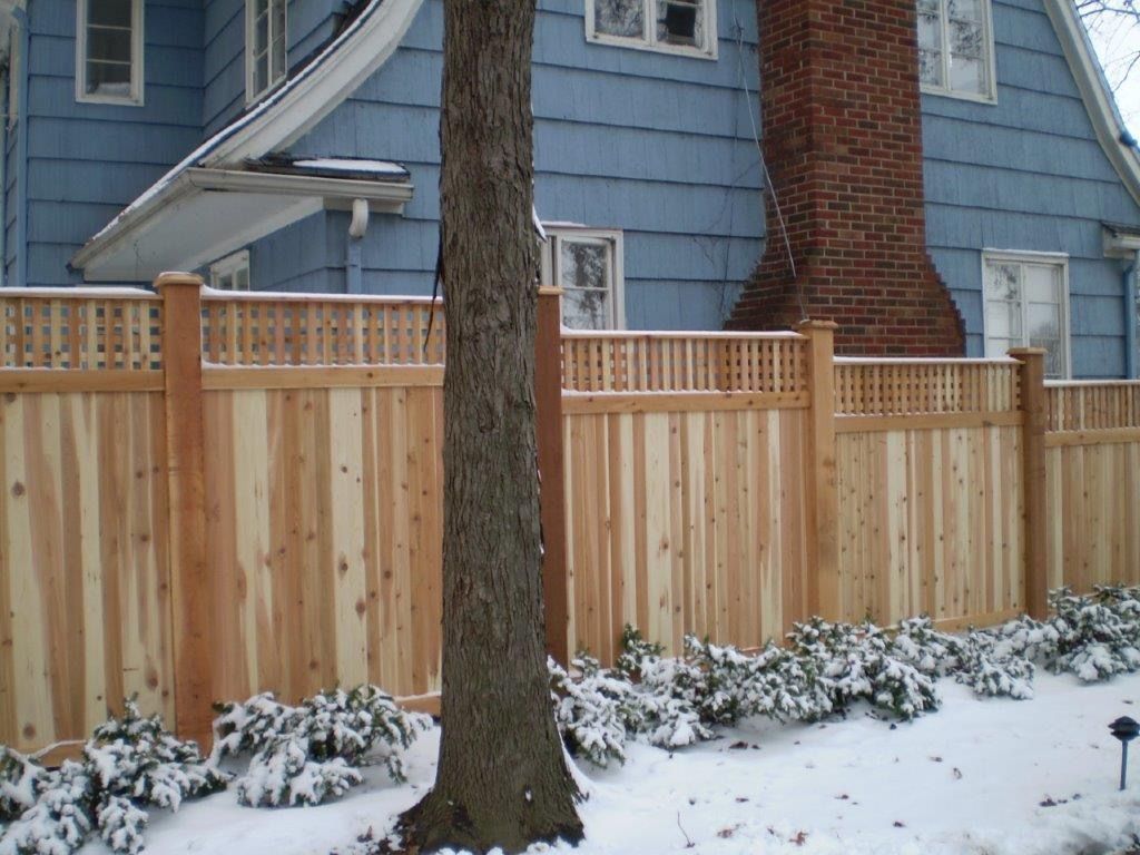 Wooden fence in front of a blue house with snow-covered bushes and a tree trunk in the foreground.
