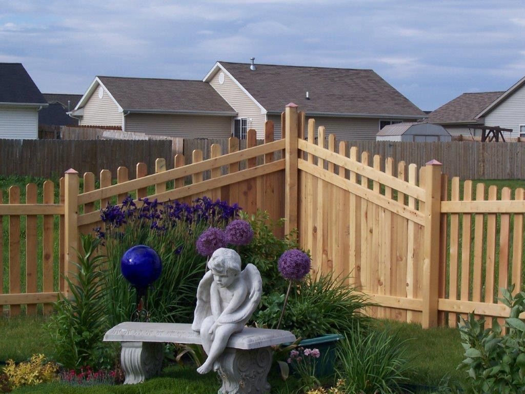 Wooden fence frames a garden with a stone bench, angel statue, and purple flowers. Houses in background.