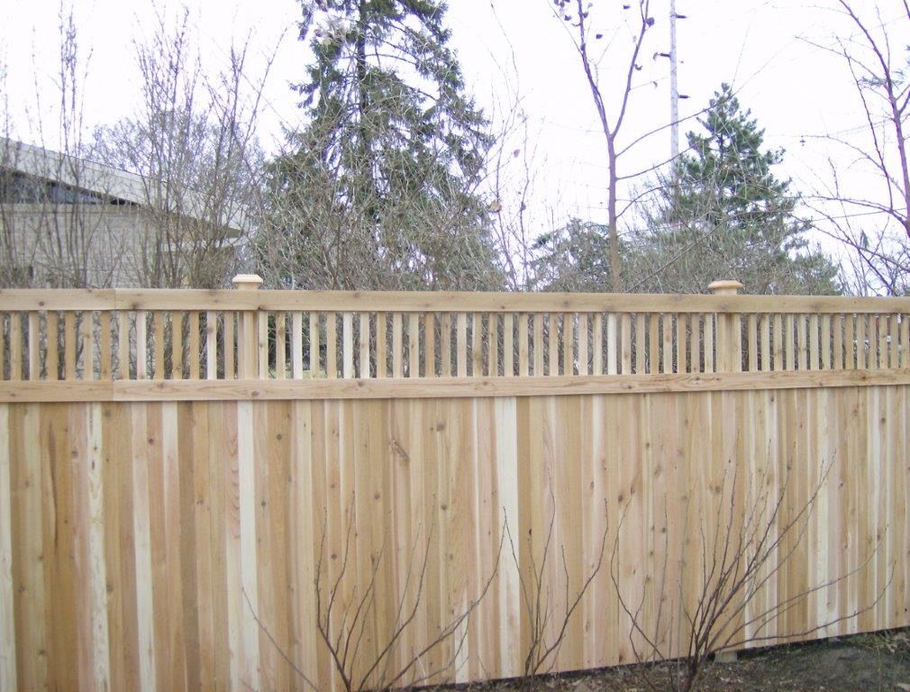 Wooden fence with vertical planks, topped with a slatted section and decorative posts, set in a yard.