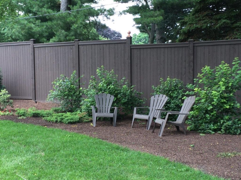 Brown privacy fence with three gray Adirondack chairs in a yard, surrounded by plants and mulch.
